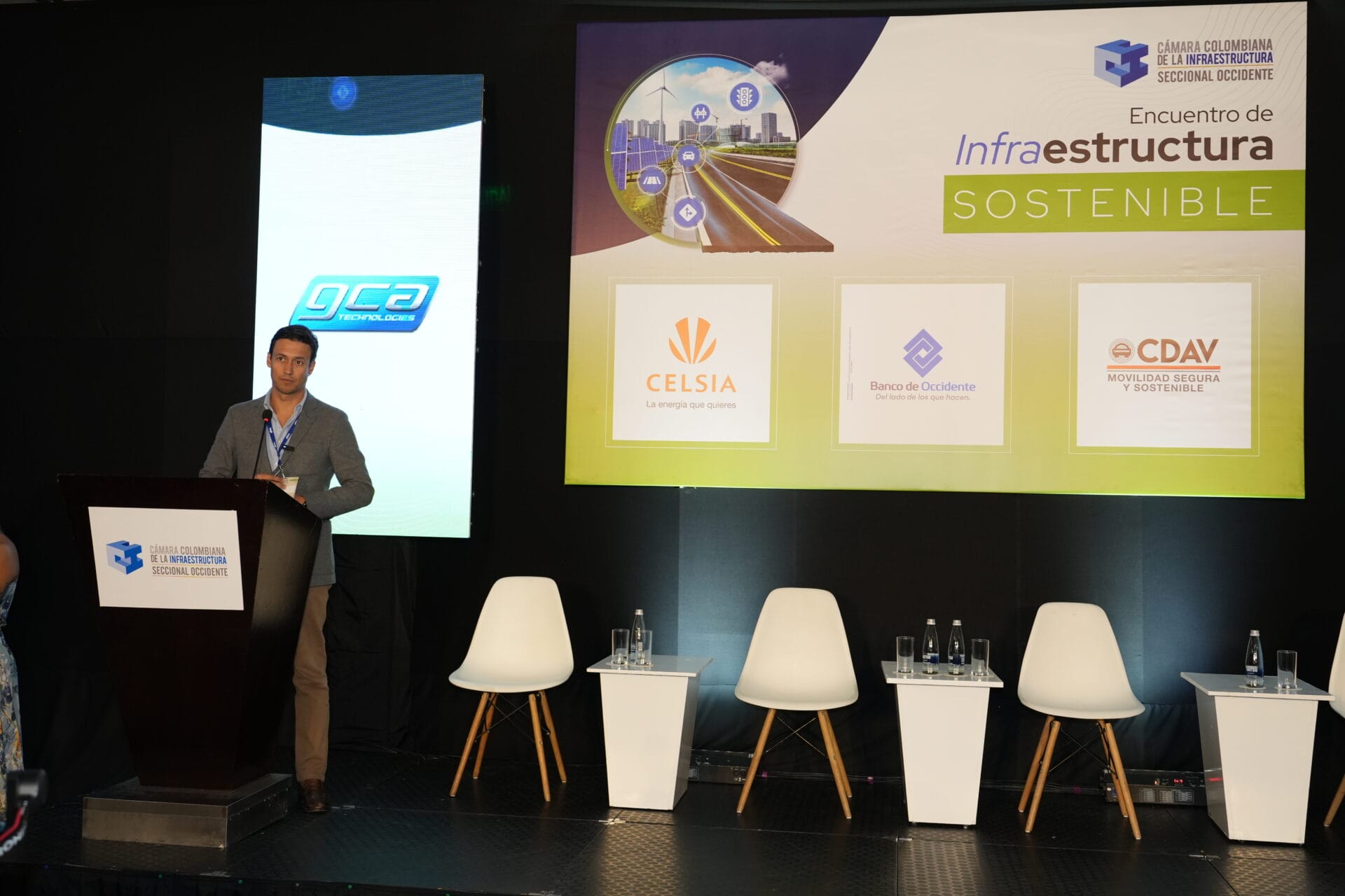 Un orador se encuentra en un podio en un escenario con tres sillas vacías y una pantalla que muestra los logotipos de Celsia, Banco de Occidente y CDAV. El evento, titulado "Infraestructura Sostenible", destaca debates sobre futuro sostenible y avances en videoanalítica inteligente.