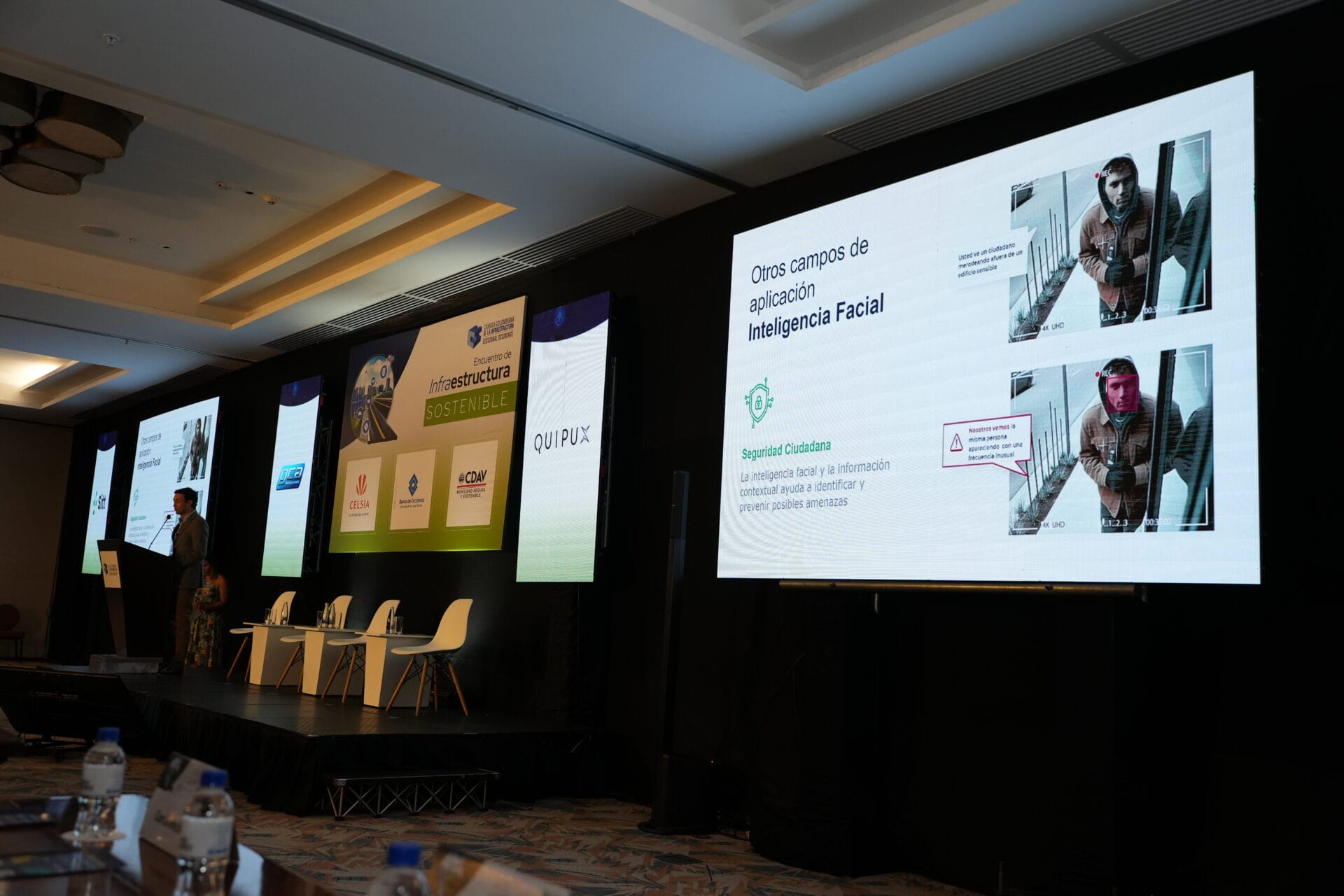 Un orador presenta una presentación de diapositivas sobre la tecnología de reconocimiento facial en una sala de conferencias moderna y sostenible con una gran pantalla que muestra diapositivas y sillas vacías al fondo. Este sistema inteligente incorpora videoanalítica para mejorar la seguridad y la eficiencia.