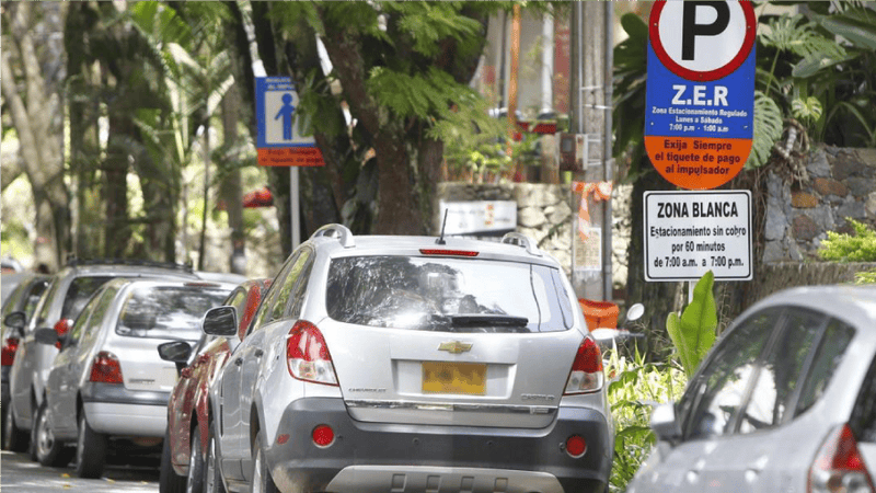Varios automóviles están estacionados a lo largo de una calle arbolada con carteles de regulación de estacionamiento visibles, incluido uno que especifica los horarios de estacionamiento pago en la Zona Blanca de 7:00 a. m. a 7:00 p. m., lo que refleja el esfuerzo que hacen las ciudades y municipios para administrar las zonas de estacionamiento regulado. y aliviar la congestión vial.