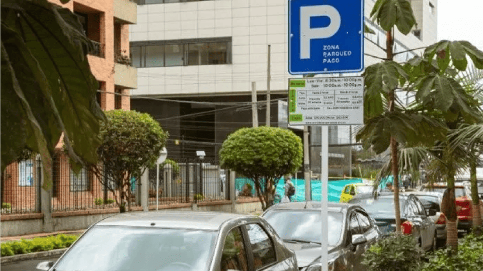 Una calle con coches aparcados junto a un cartel de "zona de aparcamiento" en español frente a un edificio. Árboles y una valla bordean la calle.